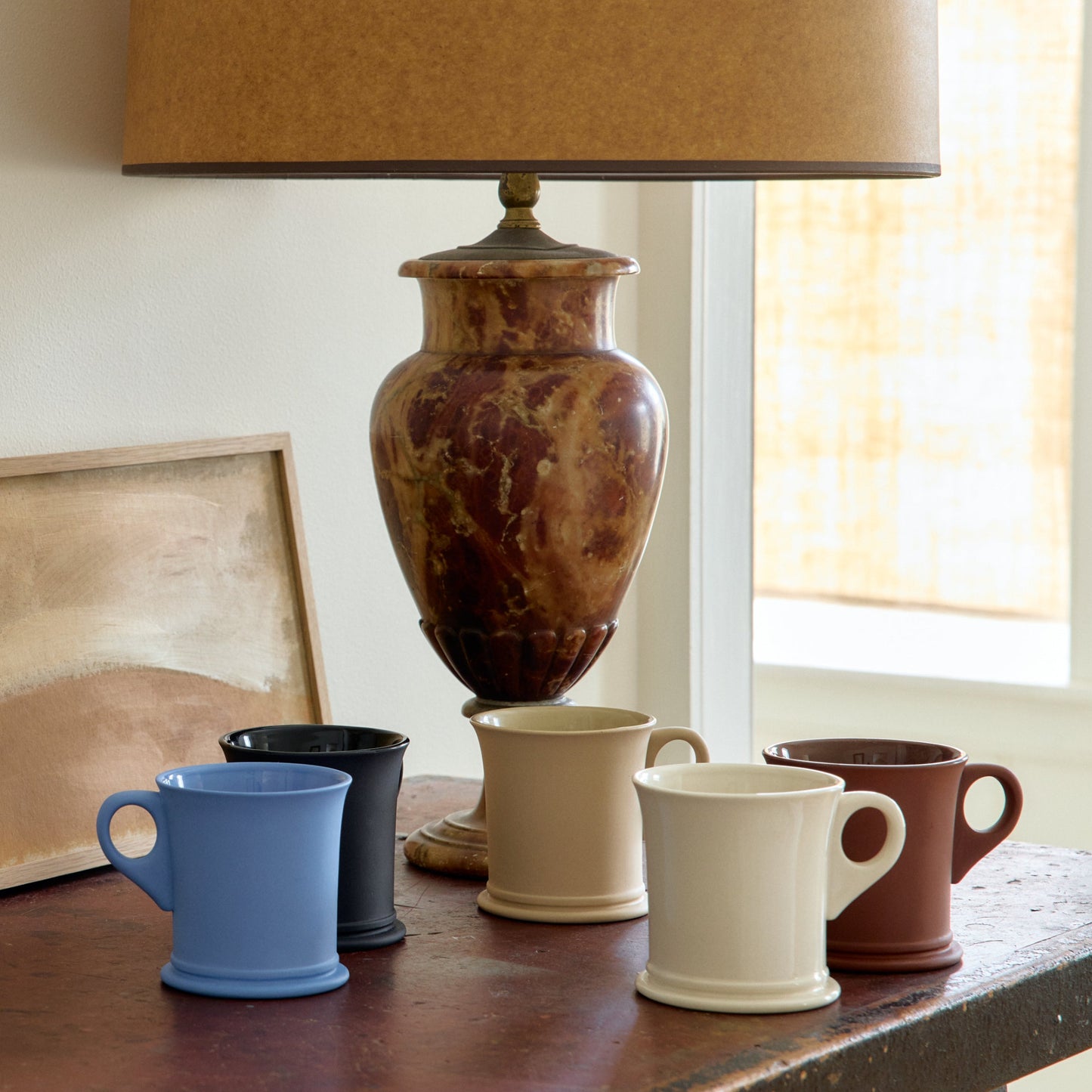 Collection of ceramic mugs on a wooden surface with a lamp and framed artwork in the background.